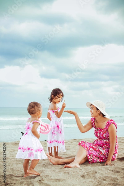 Fototapeta Asian mother and her two little children in beach dresses having fun together during vacation at a tropical beach on a summer day with blue ocean and cloudy sky on the background