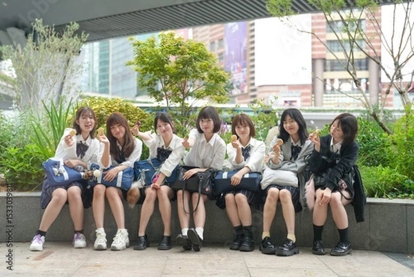 Fototapeta In early summer in Shanghai, near The Bund, Japanese high school girls in uniforms sit at a street corner, holding Chinese snacks and posing for a commemorative photo with cheerful expressions.