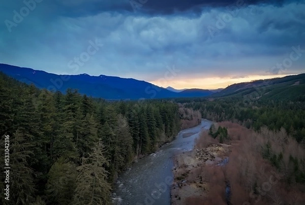 Fototapeta Aerial view of a river flowing through a forest with mountains and cloudy sky in the background
