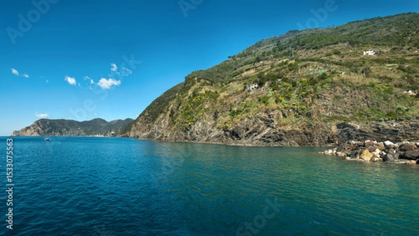 Fototapeta The coast of Cinque Terre with crystal clear water and green terraces on the slopes. A calm bay with a sailboat near the rocky shore creates an atmosphere of Italian harmony.