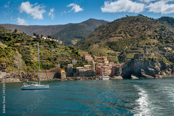 Fototapeta The seaside village of Vernazza, Cinque Terre, shines with colorful buildings, towers fortress nestled between terraced hills, sailboat on the calm blue waters of the Ligurian Sea.