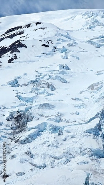 Obraz A snowy mountain slope with visible crevasses and rocky outcrops under a cloudy sky in winter
