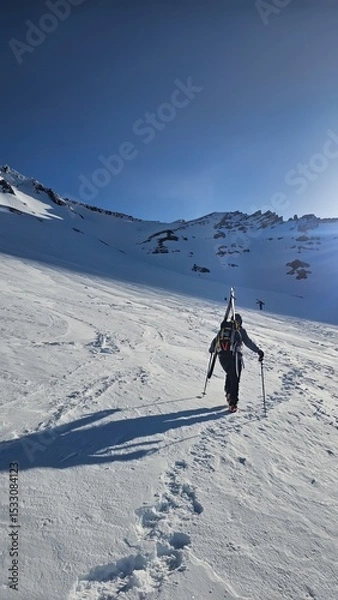 Obraz A person hiking up a snowy mountain with skis strapped to their back under a clear blue sky day
