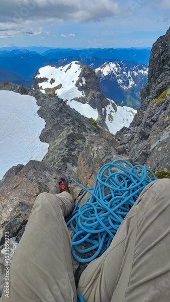 Obraz A person sitting on a rocky mountain ledge with a blue climbing rope and snowy mountain views behind