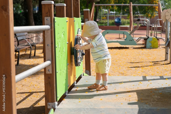 Obraz A baby boy wearing a sun hat, striped shirt, and shorts explores a playground. He is interacting with a green activity panel designed for sensory play.  Sunny summer weather