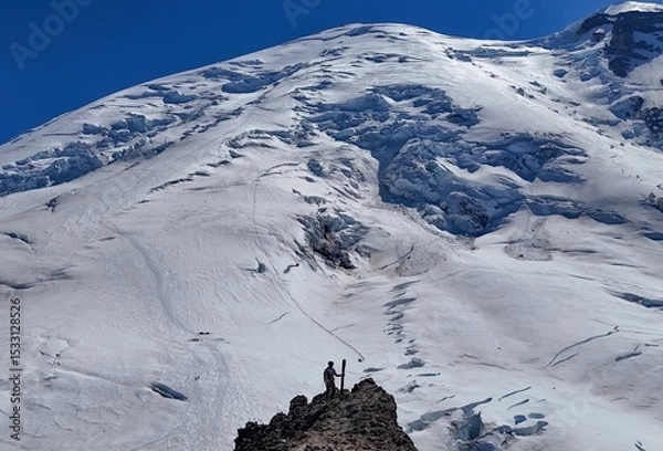 Obraz A climber stands on a rocky outcrop with skis against a backdrop of a snow covered mountain and blue sky