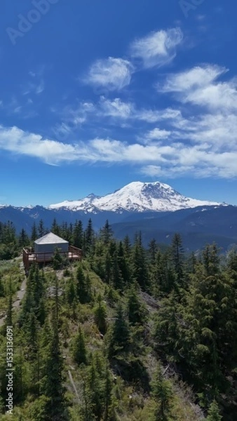Obraz Aerial view of a wooden structure on a forested hilltop with a snow capped mountain in the distance