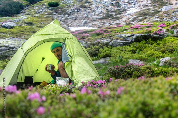 Fototapeta Camper enjoying morning coffee in green tent surrounded by pink wildflowers. Hiker relaxing at scenic mountain campsite with blooming heather.