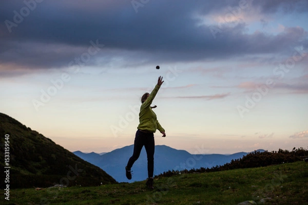 Fototapeta Hiker playing catch on mountain ridge during golden hour, with layered peaks and colorful sky creating perfect backdrop for outdoor activity.