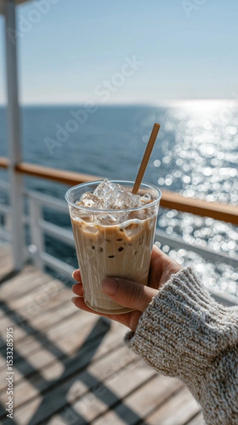 Fototapeta Hand gripping cold coffee drink with paper straw, ocean waves blurring background