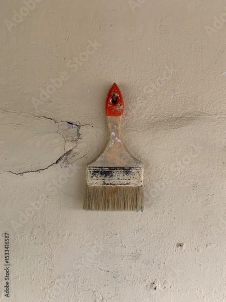 Fototapeta An old, worn paintbrush with frayed bristles leans against a damaged wall. The aged wall features cracks and a visible hole, evoking a rustic and weathered aesthetic.