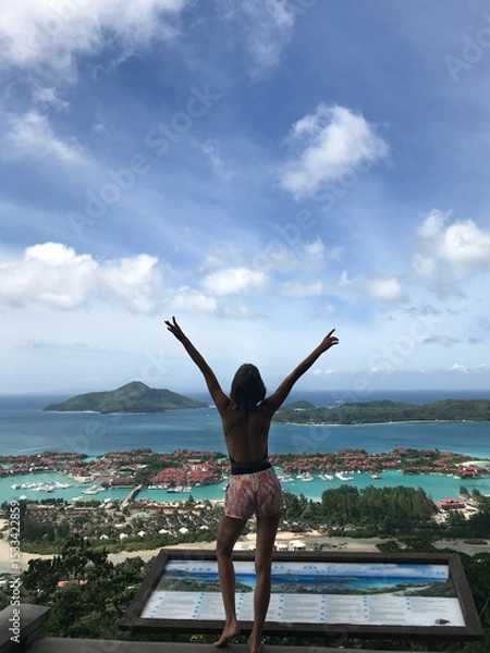 Fototapeta Young woman standing back to camera facing spectacular tropical view of island Mahe, Seychelles. Beautiful model with raised arms is enjoying traveling  on tropical destination.