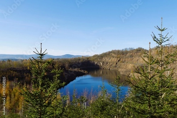 Fototapeta Scenic mountain quarry lake landscape with evergreen trees under clear blue sky, tranquil outdoor nature environment with deep water reflections surrounded by lush forest wilderness