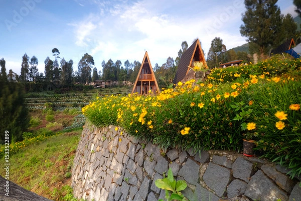 Obraz Wooden A-frame cabins situated on a vibrant flower-decorated terrace surrounded by hills under a clear blue sky, evoking tranquility, nature, and a rustic escape from urban life.
