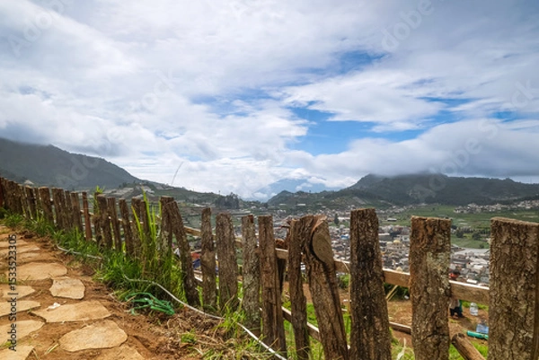 Obraz Scenic view of a rural landscape with wooden fence, mountains, and cloudy sky.
