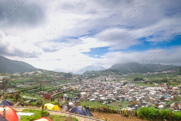 Obraz A scenic view of a town surrounded by lush green hills under a cloudy sky.