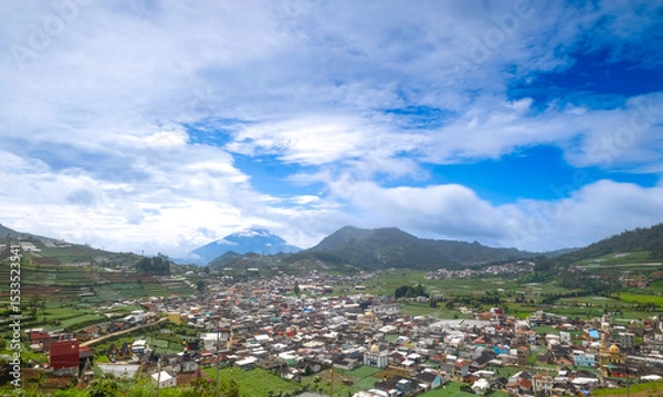 Obraz A scenic view of a town surrounded by lush green hills and mountains under a blue sky with clouds.