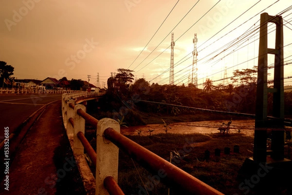 Obraz A scenic view of a bridge at sunset with power lines and a river below, surrounded by trees and a warm orange sky.