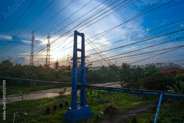 Obraz A blue tower structure with power lines against a sunrise sky, surrounded by greenery.