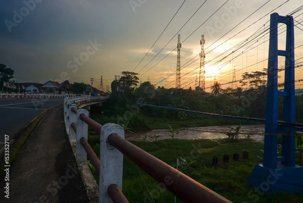 Obraz A serene sunset over a rural bridge with power lines and lush greenery.