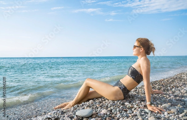 Fototapeta A young girl in a bikini is sunbathing, sitting at the edge of the sea on a pebble beach against a background of turquoise sea and blue sky. Side view