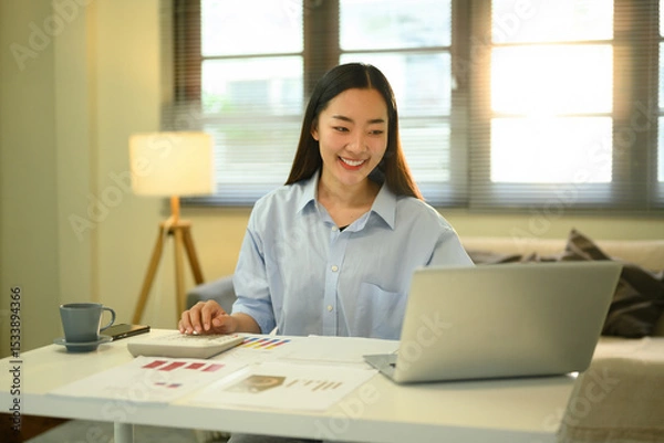 Fototapeta Cheerful female professional analyzing reports at a bright, modern home office desk with digital tools and documents