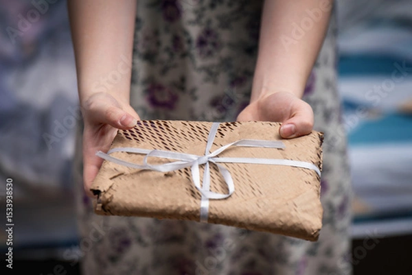 Fototapeta Young woman offering a gift-wrapped book in craft perforated paper with a bow, showcasing thoughtful presentation