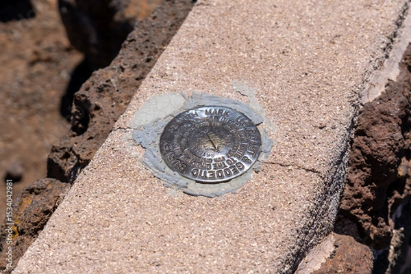 Fototapeta Geodetic survey marker embedded in concrete, surrounded by dark rocks, indicating a precise location.