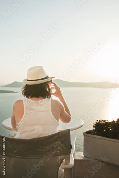 Fototapeta A woman enjoys a scenic sunset view over the Aegean Sea from a Santorini terrace with a glass of wine.