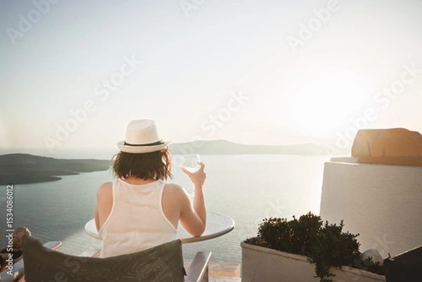 Fototapeta A woman enjoys a scenic sunset view over the Aegean Sea from a Santorini terrace with a glass of wine.