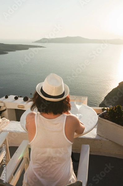 Fototapeta A woman enjoys a scenic sunset view over the Aegean Sea from a Santorini terrace with a glass of wine.
