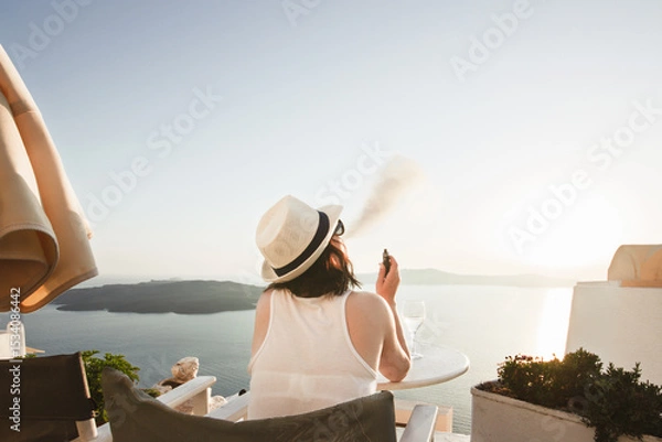 Fototapeta A woman enjoys a scenic sunset view over the Aegean Sea from a Santorini terrace with a glass of wine and a vape.