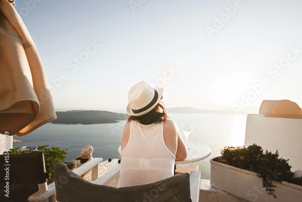 Fototapeta A woman enjoys a scenic sunset view over the Aegean Sea from a Santorini terrace with a glass of wine and a vape.