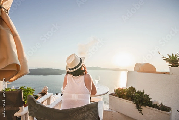 Fototapeta A woman enjoys a scenic sunset view over the Aegean Sea from a Santorini terrace with a glass of wine and a vape.