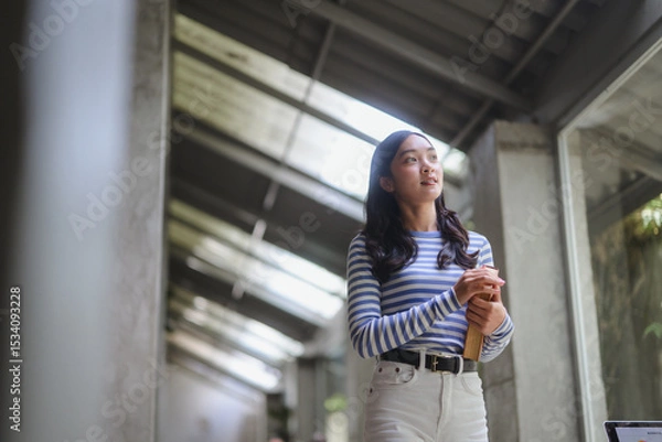 Fototapeta Young student walking in university corridor holding a book and looking up