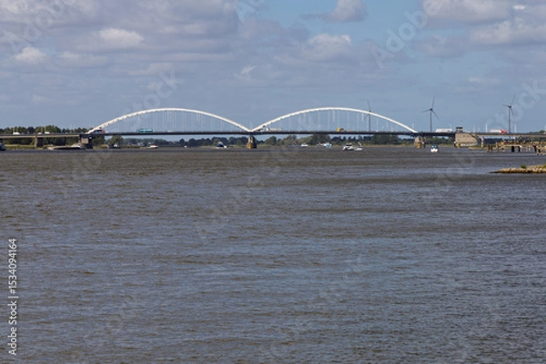 Obraz The Merwedebrug Gorinchem (Merwede Bridge) arches gracefully over a wide river, bustling with boats and traffic, under a bright, cloudy Dutch sky.