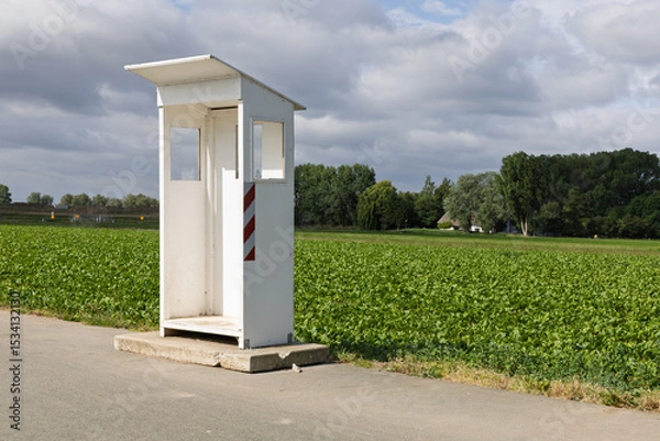Obraz An empty white booth, a guard booth, stands by a rural road with a vibrant green field and cloudy sky in the background.