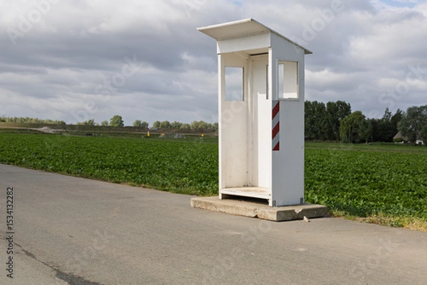 Obraz An isolated white booth, a guard shack, stands by a rural road next to a vibrant green field under a cloudy sky.