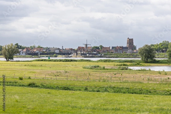 Obraz A beautiful panoramic view of the historic town of Woudrichem, Netherlands, featuring its iconic church and windmill, nestled by a wide river and lush green fields. A classic Dutch landscape.