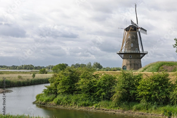 Obraz This scenic shot captures a classic Dutch windmill Gorinchem. standing tall against a cloudy sky, overlooking a tranquil waterway and lush green landscape. A symbol of the Netherlands.
