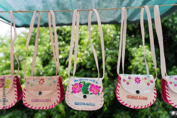 Fototapeta Handcrafted souvenir leather bags with floral embroidery and "Guadalajara" text hanging on display at a local market in Jalisco, Mexico. Language translation: I thought of you in Guadalajara