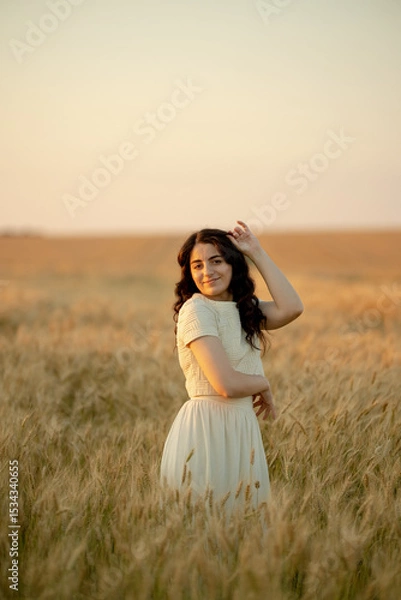 Fototapeta A young brunette walks in wheat fields at sunset in summer. Growing organic wheat and barley.
