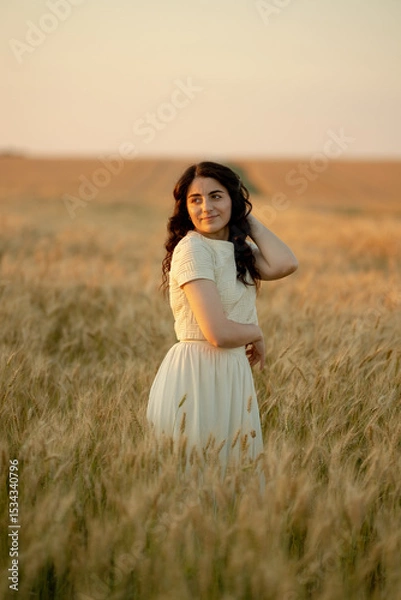 Fototapeta A young brunette walks in wheat fields at sunset in summer. Growing organic wheat and barley.