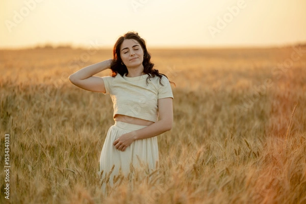 Fototapeta A young brunette walks in wheat fields at sunset in summer. Growing organic wheat and barley.