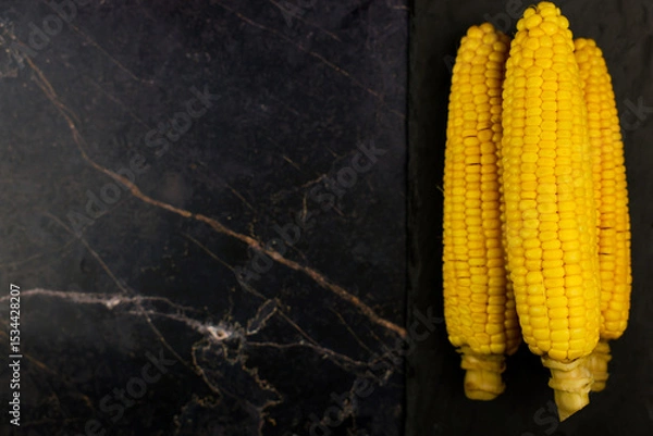 Fototapeta Yellow ripe peeled ears of corn with large grains lie on a black stone plank on the side on a dark background with space for text. The concept of healthy nutrition, natural iron, corn cultivation
