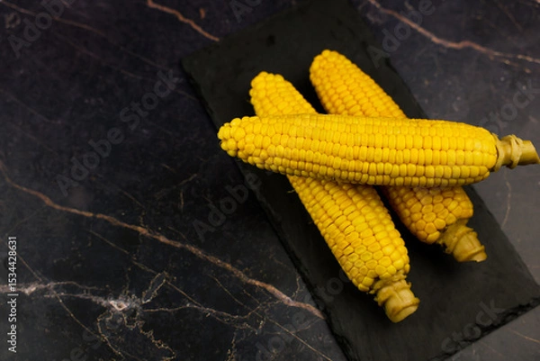 Fototapeta Yellow ripe peeled ears of corn with large grains and stem stem lie on a black stone plank on the side on a dark background with space for text. The concept of healthy nutrition, natural iron, corn