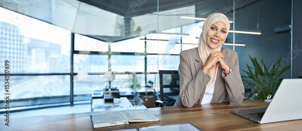Fototapeta Portrait young muslim specialist business lady working on laptop pc in office smiling at camera. Middle eastern indian woman in hijab using computer technology for work online. Banner, copy space