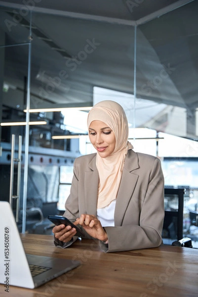 Fototapeta Middle Eastern muslim businesswoman manager ceo using cell phone mobile application. Smiling young indian woman in hijab holding smartphone sitting in office working online at desk on gadget. Vertical