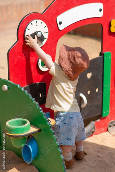 Obraz Baby boy dressed with summer cloves and hat is playing outside on the street, touching and exploring playground for little ones. The child is reaching up to interact with a red play structure.