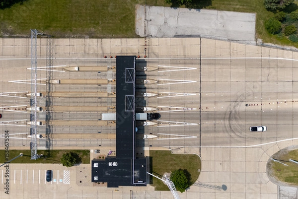 Fototapeta Top-down aerial view of a highway toll plaza with multiple lanes, a central toll booth building, and vehicles including a semi-truck passing through under bright daylight in a suburban area.